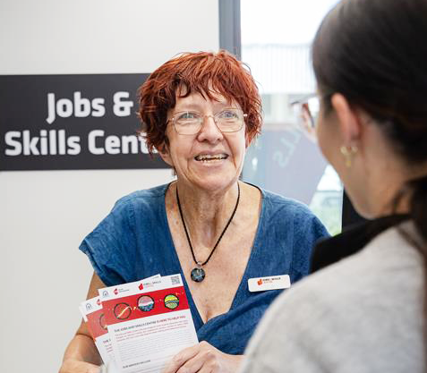 a person holding pamphlets speaks with another person in front of a “Jobs & Skills Centre” sign