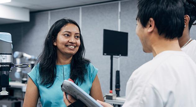 TAFE lecturer speaking with students in a technology training laboratory, with robotic automation equipment and digital tools visible in the workspace.