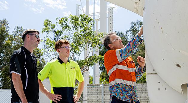 TAFE lecturer demonstrating maintenance work on a wind turbine while two students observe at a renewable energy training facility.
