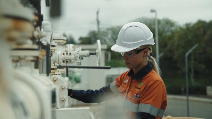 Person in high visibility clothing and a white hard hat inspecting pipes in a yard.